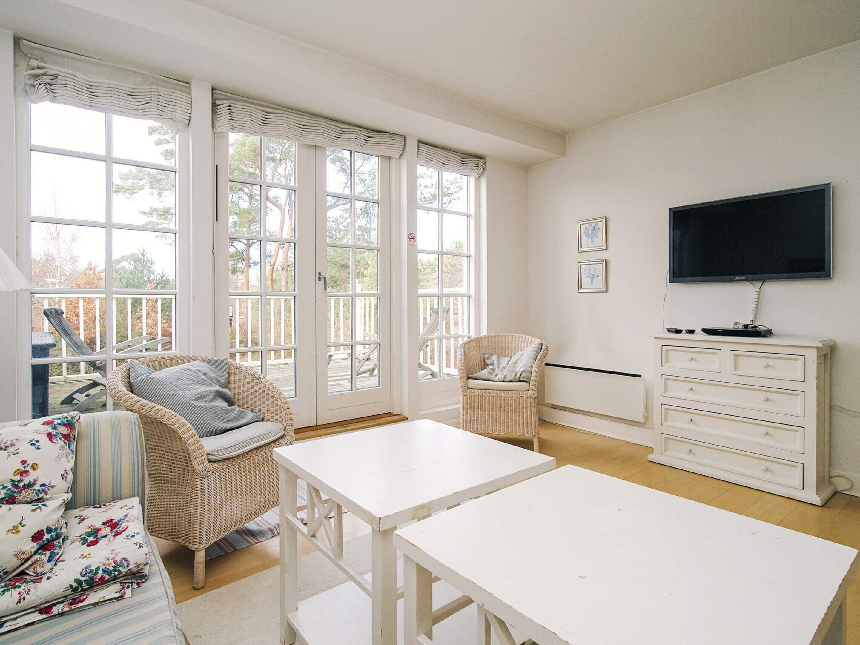 Una habitación luminosa con ventanas amplias, muebles de mimbre, una mesa blanca y un televisor en la pared.