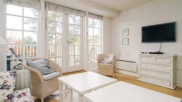 Una habitación luminosa con ventanas amplias, muebles de mimbre, una mesa blanca y un televisor en la pared.