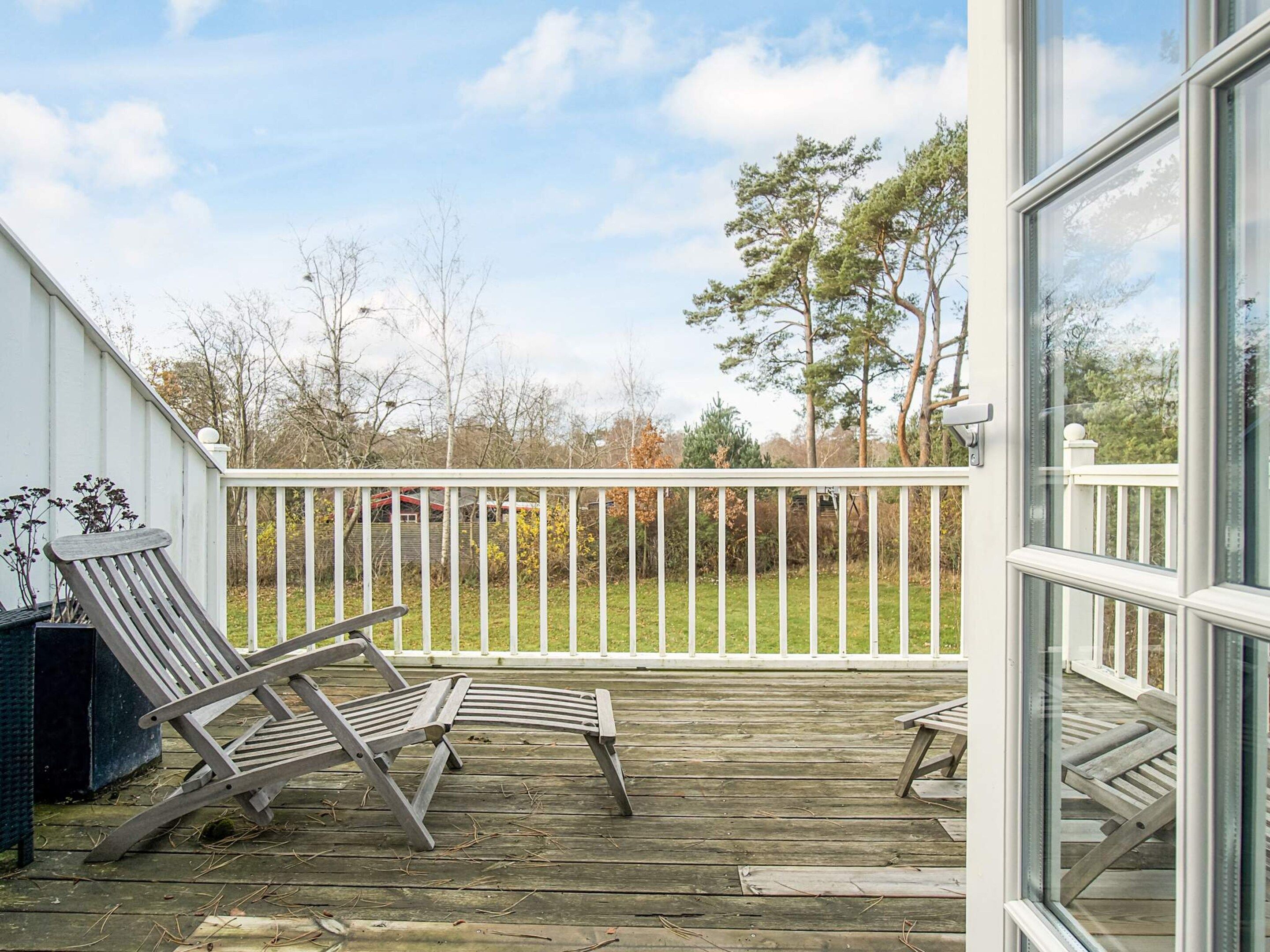 Une terrasse en bois avec deux chaises longues, une clôture blanche et, en arrière-plan, une vue sur des arbres et un bâtiment.