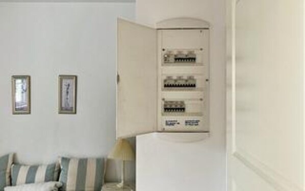 Quarto com parede branca, quadro emoldurado, luminária e painel elétrico.