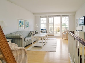 Ein helles Wohnzimmer mit einem gestreiften Sofa, einem weißen Tisch, Korbstühlen und Blick durch eine Glastür auf eine Terrasse.