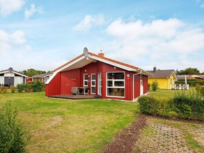 A red building with a porch and a small table, surrounded by a grassy yard and a paved walkway. - Holiday Home in GrÃ¶mmitz (Groemitz)