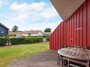 A wooden deck with a table and chairs, a red building, and a grassy yard with a hedge. - Holiday Home in GrÃ¶mmitz (Groemitz)