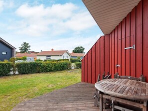 A wooden deck with a table and chairs, a red building, and a grassy yard with a hedge.
