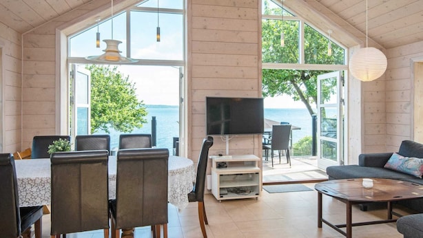 A spacious living room with a large window overlooking the sea, a dining area with chairs and a table, and a cozy seating area with a sofa. - Panoramic Retreat by Water (Borkop)