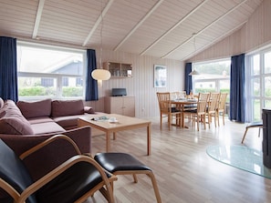 A spacious living area with a sofa, dining table, and chairs, large windows with curtains, and a wooden ceiling. - Holiday Home in Grömmitz (Groemitz)