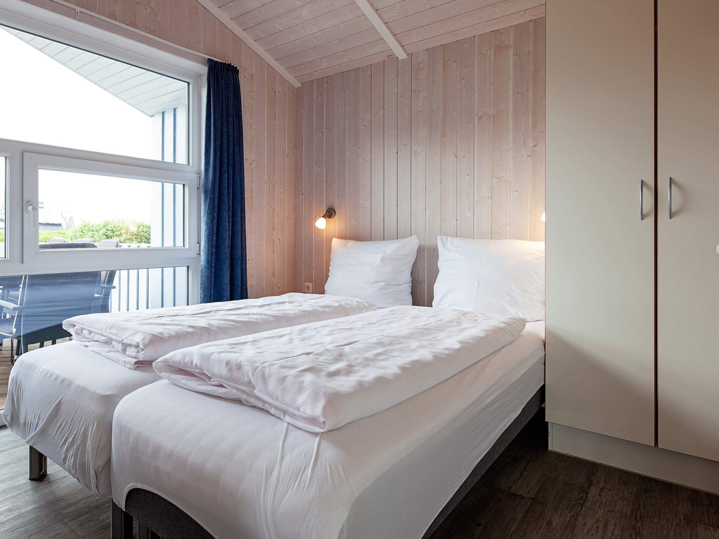 A double bed with white linens, a wooden wardrobe, and a window with curtains.