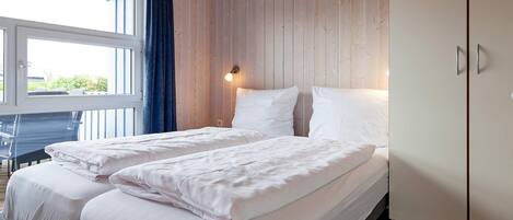 A double bed with white linens, a wooden wardrobe, and a window with curtains.