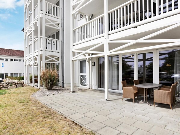 Un bâtiment moderne à l’extérieur blanc, doté d’un espace extérieur abrité avec des meubles en osier, et d’un mur en pierre agrémenté d’une petite plante.