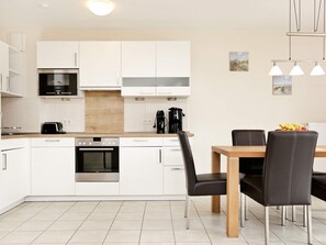A modern kitchen with white cabinets, a built-in oven, a microwave, and a coffee maker. There is a dining table with chairs and a fruit bowl on the table. - Holiday Home in Grossenbrode (Grossenbrode)