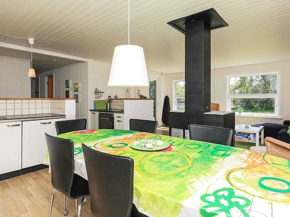 A dining area with a table covered in a colorful tablecloth, black chairs, a hanging light fixture, and a view of the kitchen and garden through a window.