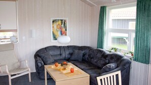 En hyggelig stue med en mørk lædersofa, et træbord og en hvid lænestol. Et maleri hænger på væggen, og et vindue med grønne gardiner er synligt.