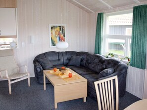 A cozy living room with a dark leather sofa, a wooden coffee table, and a white armchair. A painting hangs on the wall, and a window with green curtains is visible. - Holiday Home in Otterndorf (Otterndorf)
