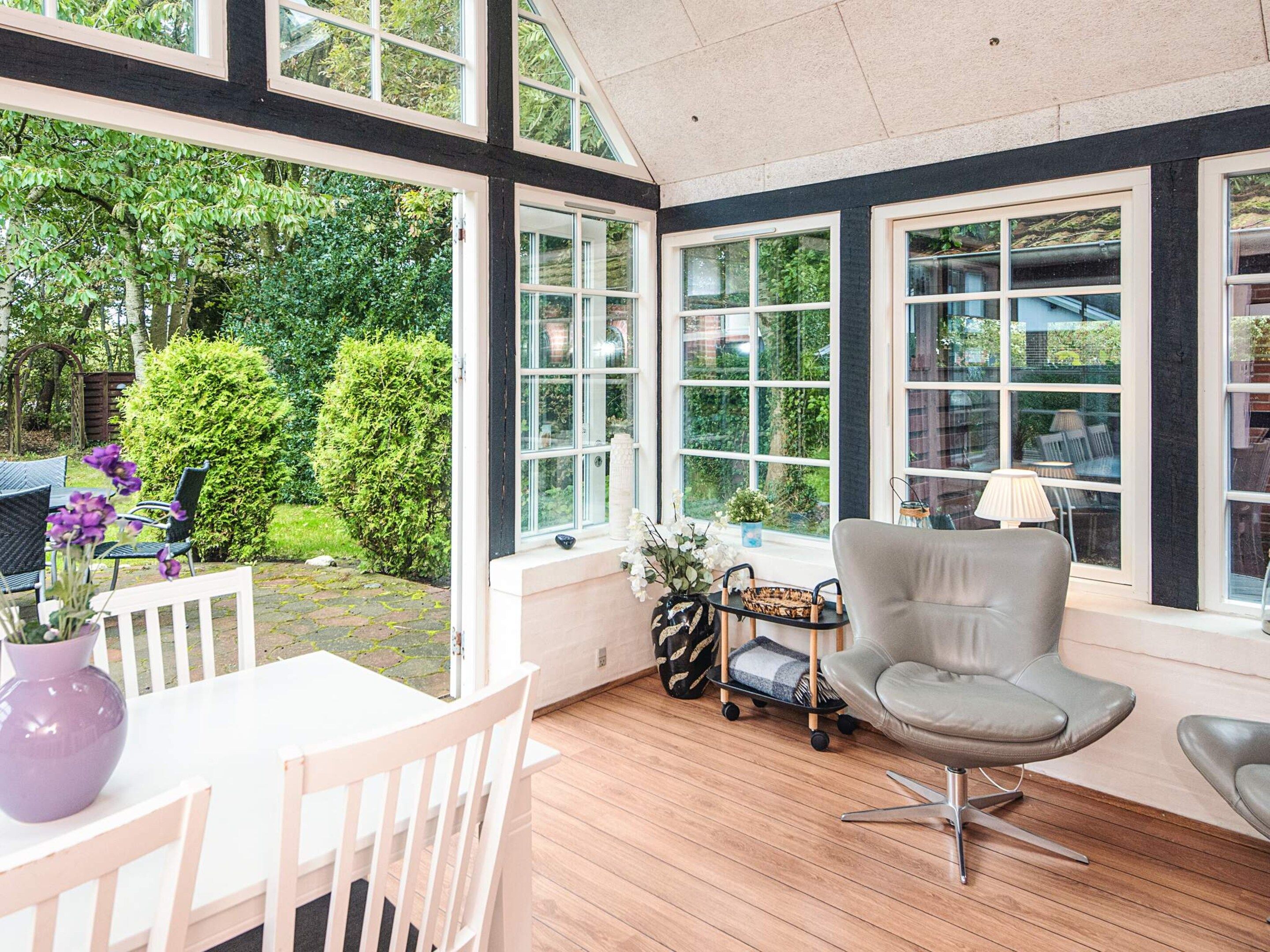 A sunlit room with a view of greenery, a white table with chairs, and a grey chair.