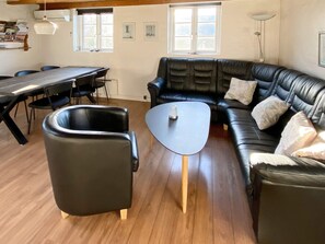 Una sala de estar con sofá de cuero negro, una mesa redonda, una mesa de comedor con sillas y un ventanal que proporciona luz natural.