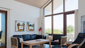 Een moderne woonkamer met een bank, een houten salontafel en twee fauteuils. Er zijn grote ramen met uitzicht op groen.