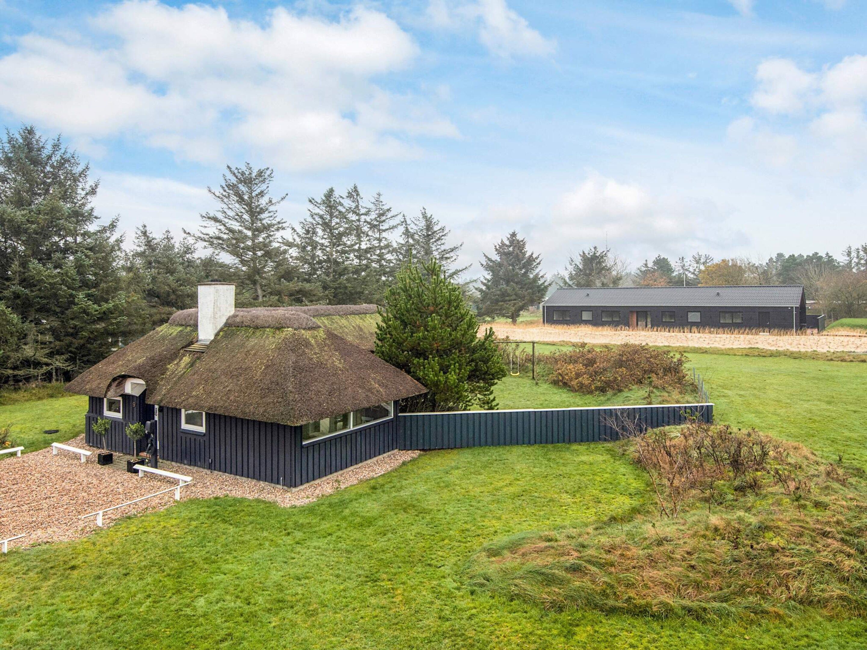 A thatched-roof house with a dark exterior, surrounded by a grassy field and trees.