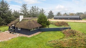 Une maison à toit de chaume, à l’extérieur sombre, entourée d’un champ herbeux et d’arbres.