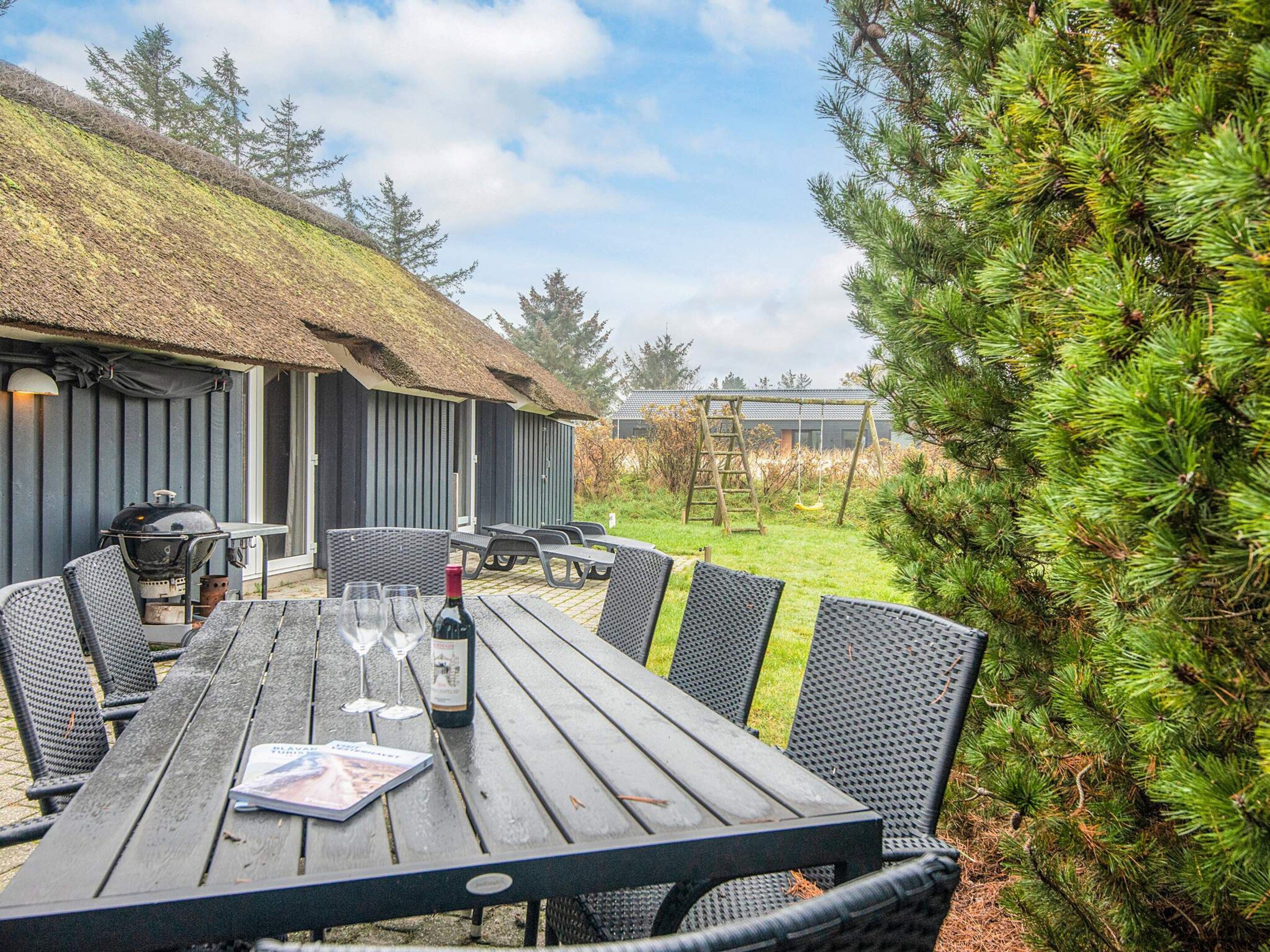 An outdoor dining area with a wooden table and chairs, a grill, and a thatched-roof building in the background.