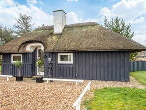 Een rieten dak huis met een donkere gevel, een schoorsteen en een klein terras met een bankje.