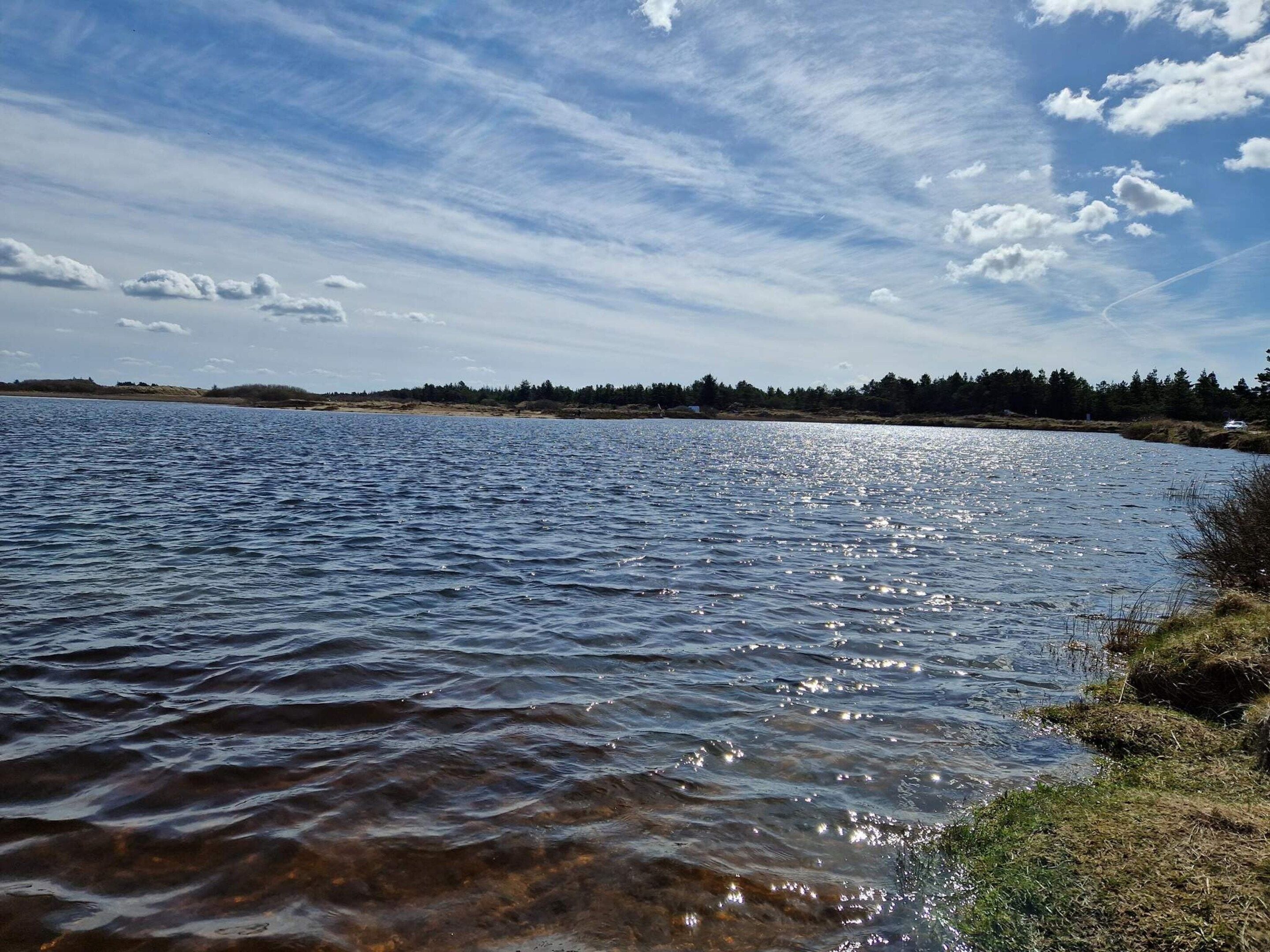 A calm lake with clear water reflecting the sky, surrounded by a forested shoreline.