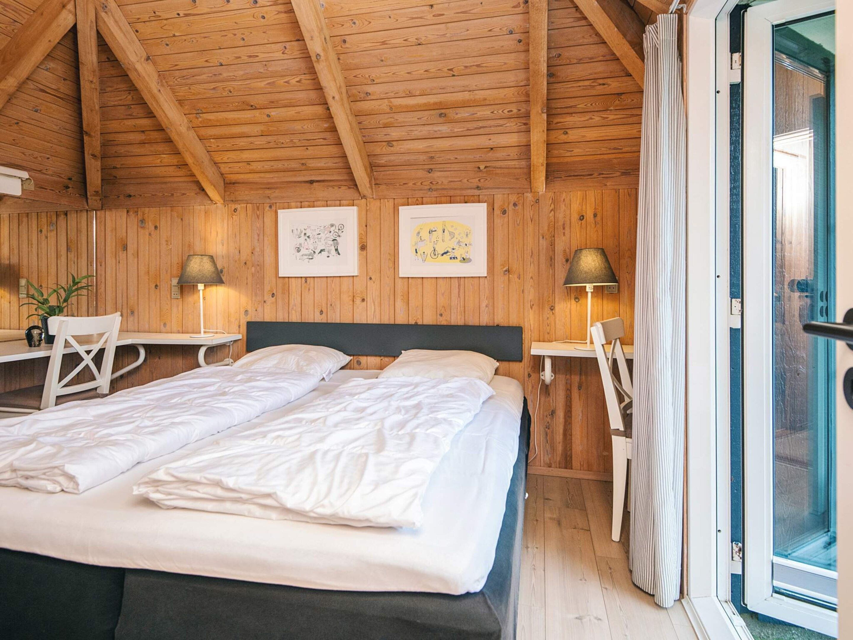 A cozy bedroom with a wooden ceiling, a bed with white linens, two bedside tables with lamps, and a window with curtains.