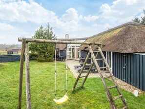 A wooden swing set in a backyard with a thatched-roof building and a deck. - Beautiful Holiday Home in Vejers Strand with Hot Tub (Vejers Strand)