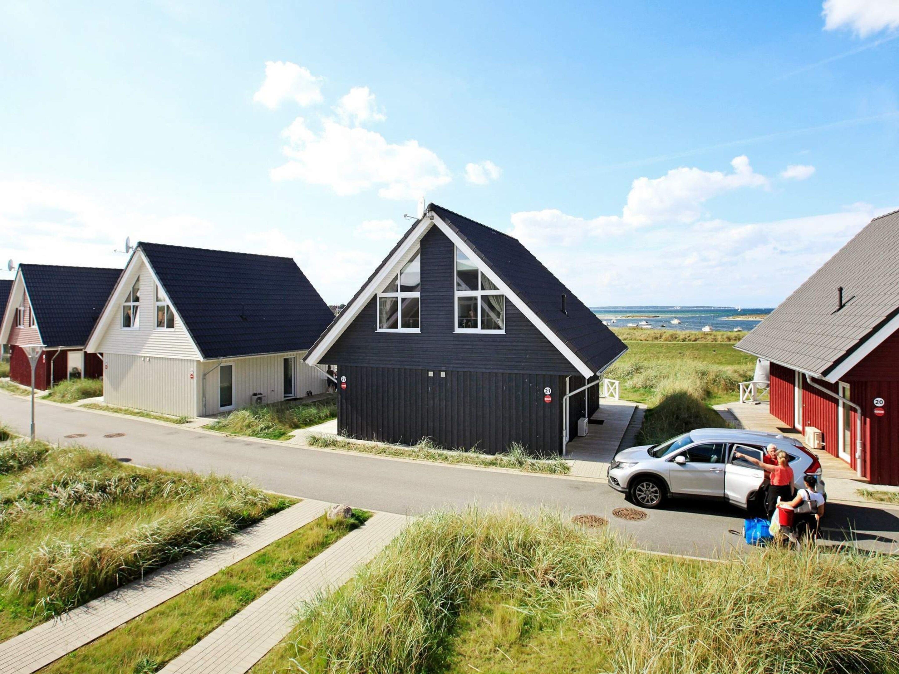 A row of modern houses with a paved road in front, a car parked on the side, and a clear view of the sea in the background.