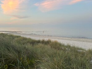 A beach with a person walking, grassy dunes, and a calm sea. - 8 Person Holiday Home in Hadsund-by Traum (Hadsund)