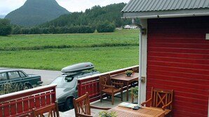 A wooden deck with outdoor seating, a red building, and a view of a green field and mountain. - Holiday Home in Isfjorden (Rauma)