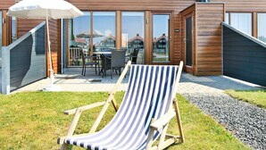 A modern building with a wooden facade, a covered patio with outdoor seating, and a grassy area with a striped lounge chair. - Bright Apartment in Bogense Denmark With Terrace (Bogense)