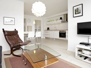 Una sala de estar moderna con una mesa de centro de vidrio, un sillón reclinable de cuero, armarios blancos, una zona de cocina y un televisor de pantalla plana.