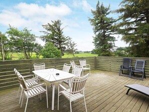 An outdoor patio with a table and chairs, surrounded by a wooden deck and a view of a green field and trees.