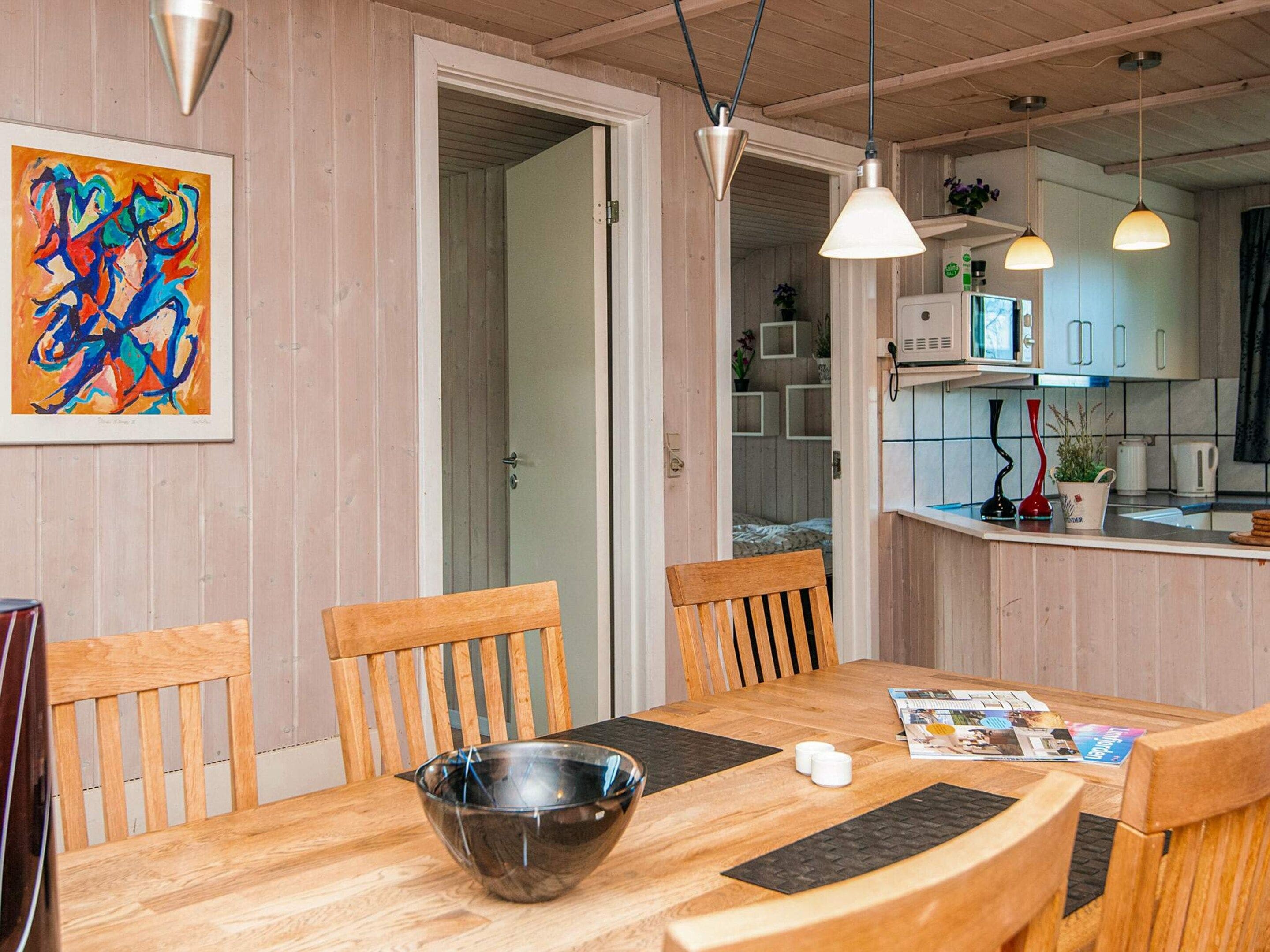 A dining area with a wooden table, chairs, a bowl, and a magazine. There is a kitchen in the background with cabinets and a microwave.