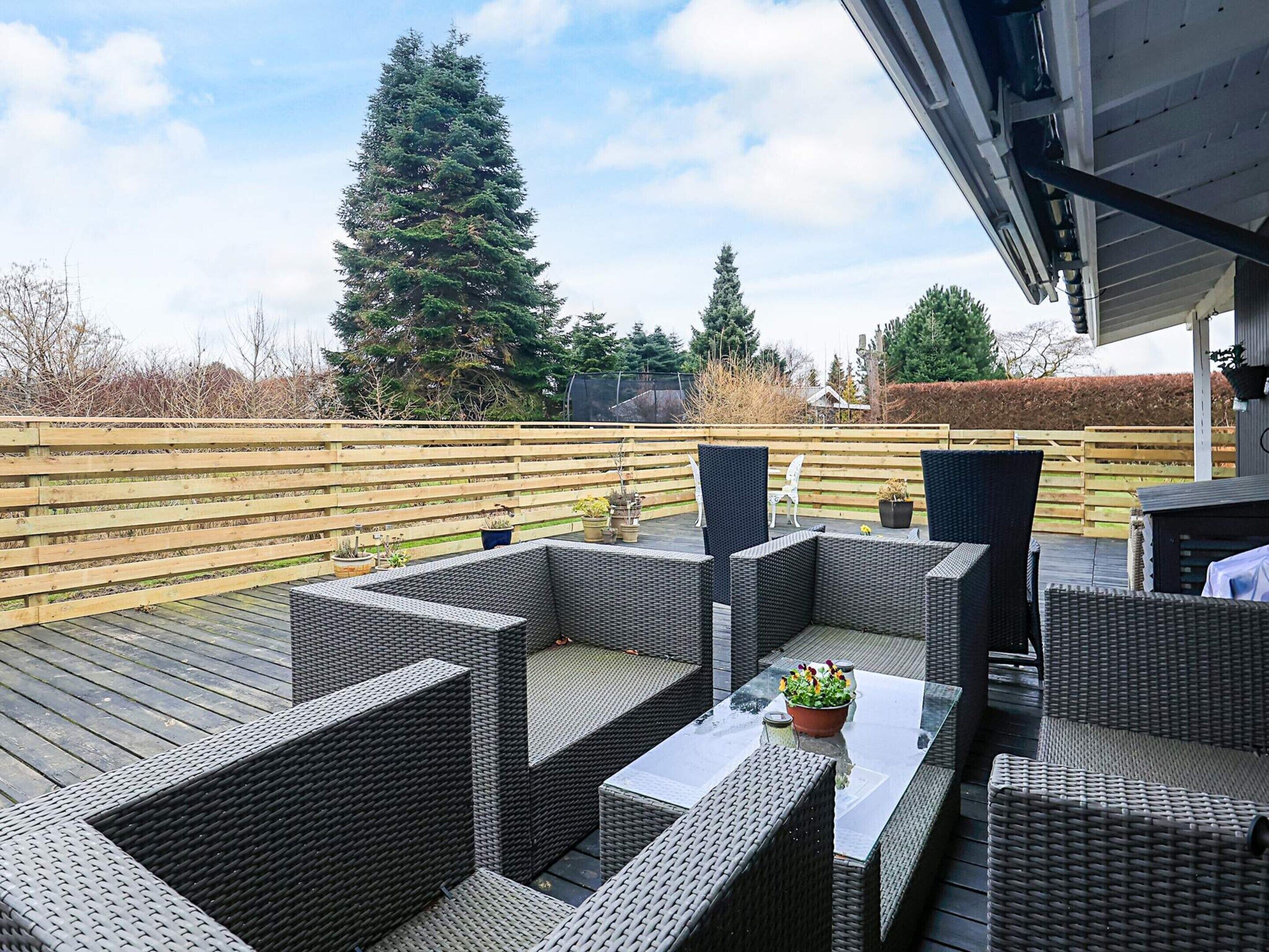 Un espace extérieur abrité, aménagé avec des meubles en osier et une table, entouré d’une terrasse en bois et d’une clôture en bois.