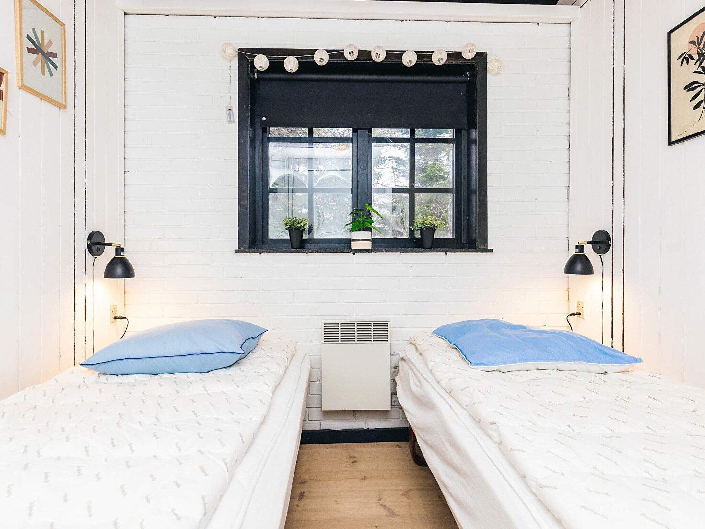 A room with two beds, a window with plants, and two wall-mounted lamps.