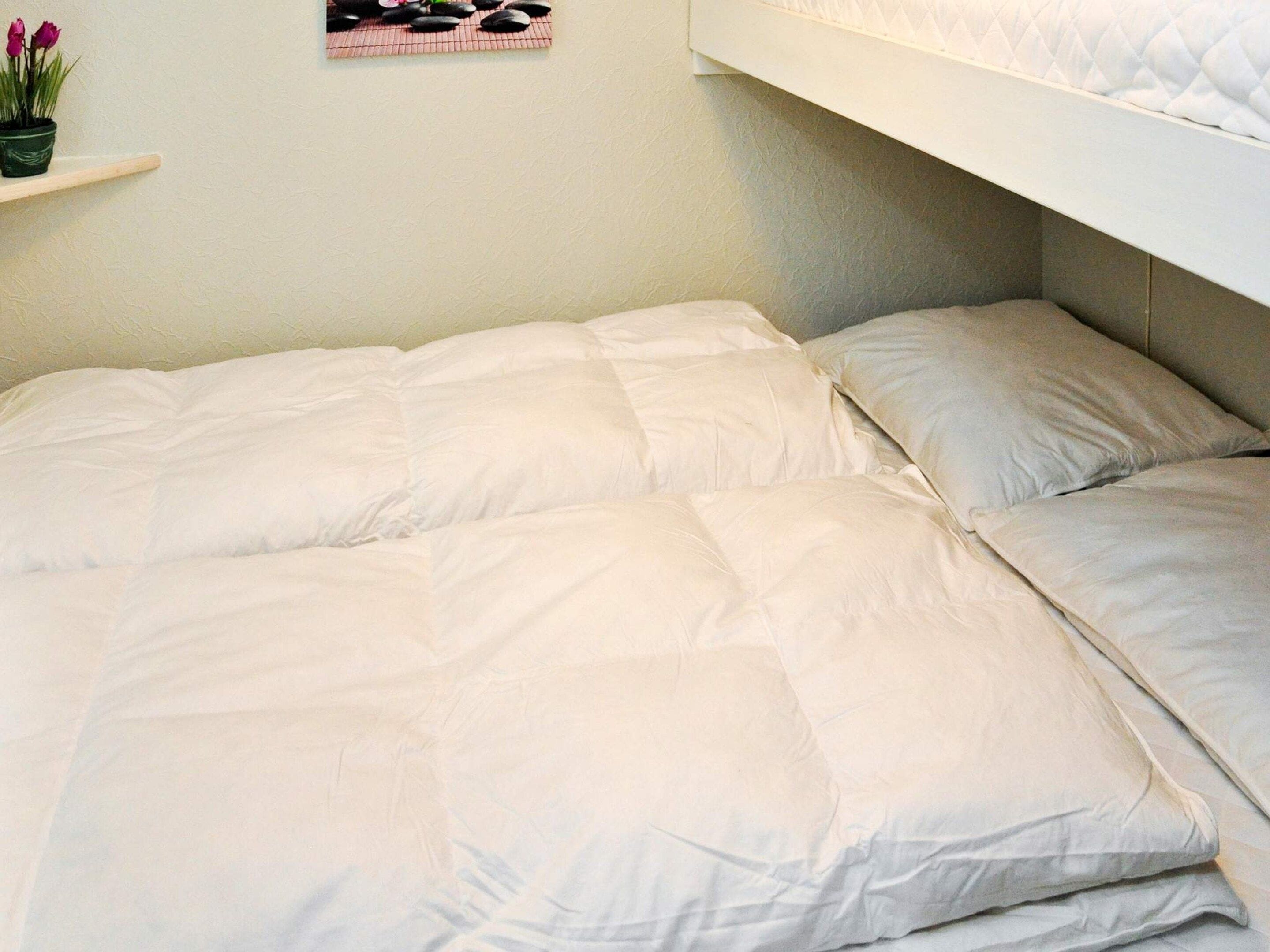 Un lit avec une couette blanche matelassée et deux oreillers, une petite étagère avec une plante en pot et un tableau accroché au mur.
