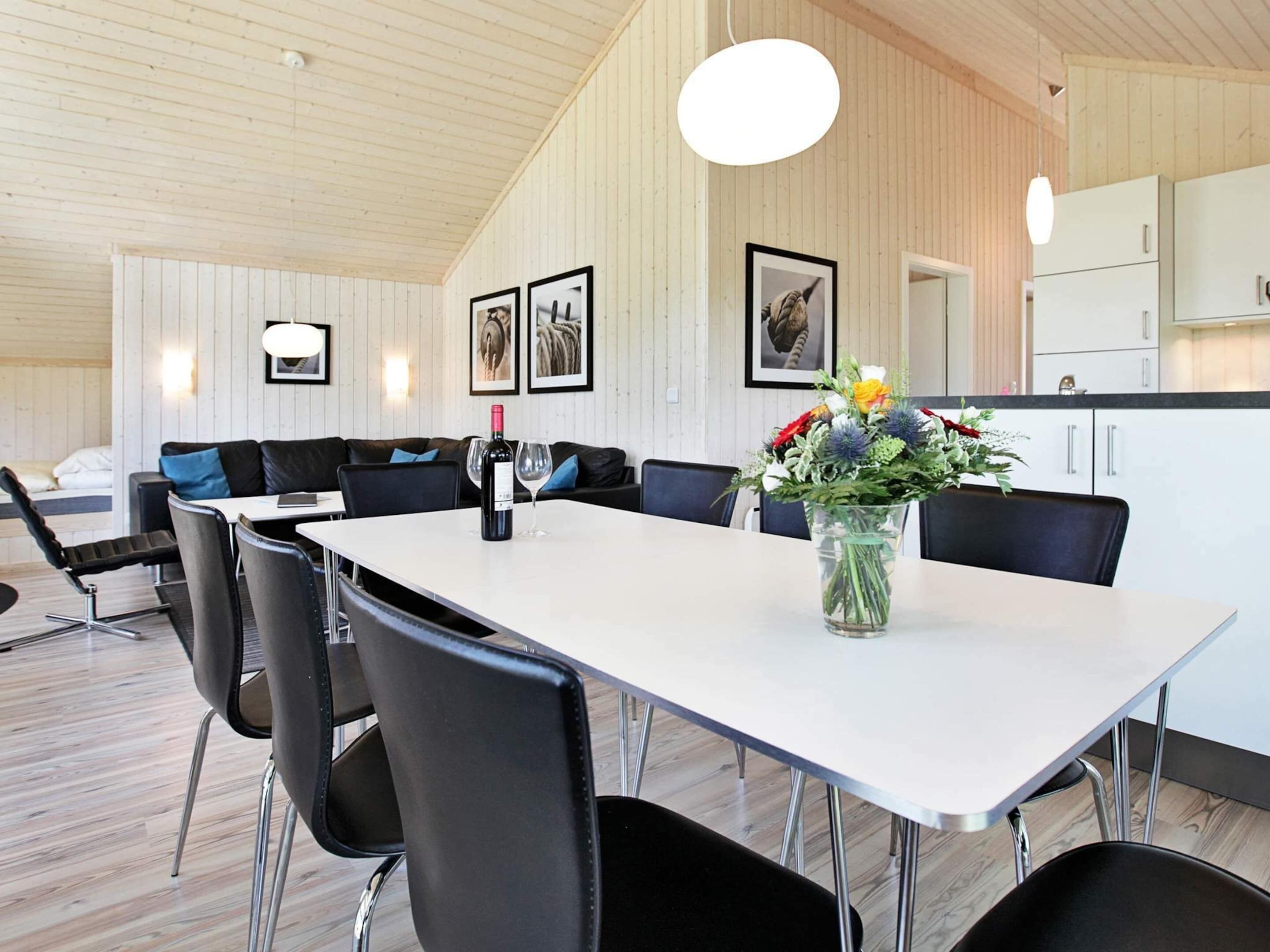 Un espace repas moderne avec une table blanche, des chaises noires et un vase de fleurs. On aperçoit un canapé et un lit à l’arrière-plan.
