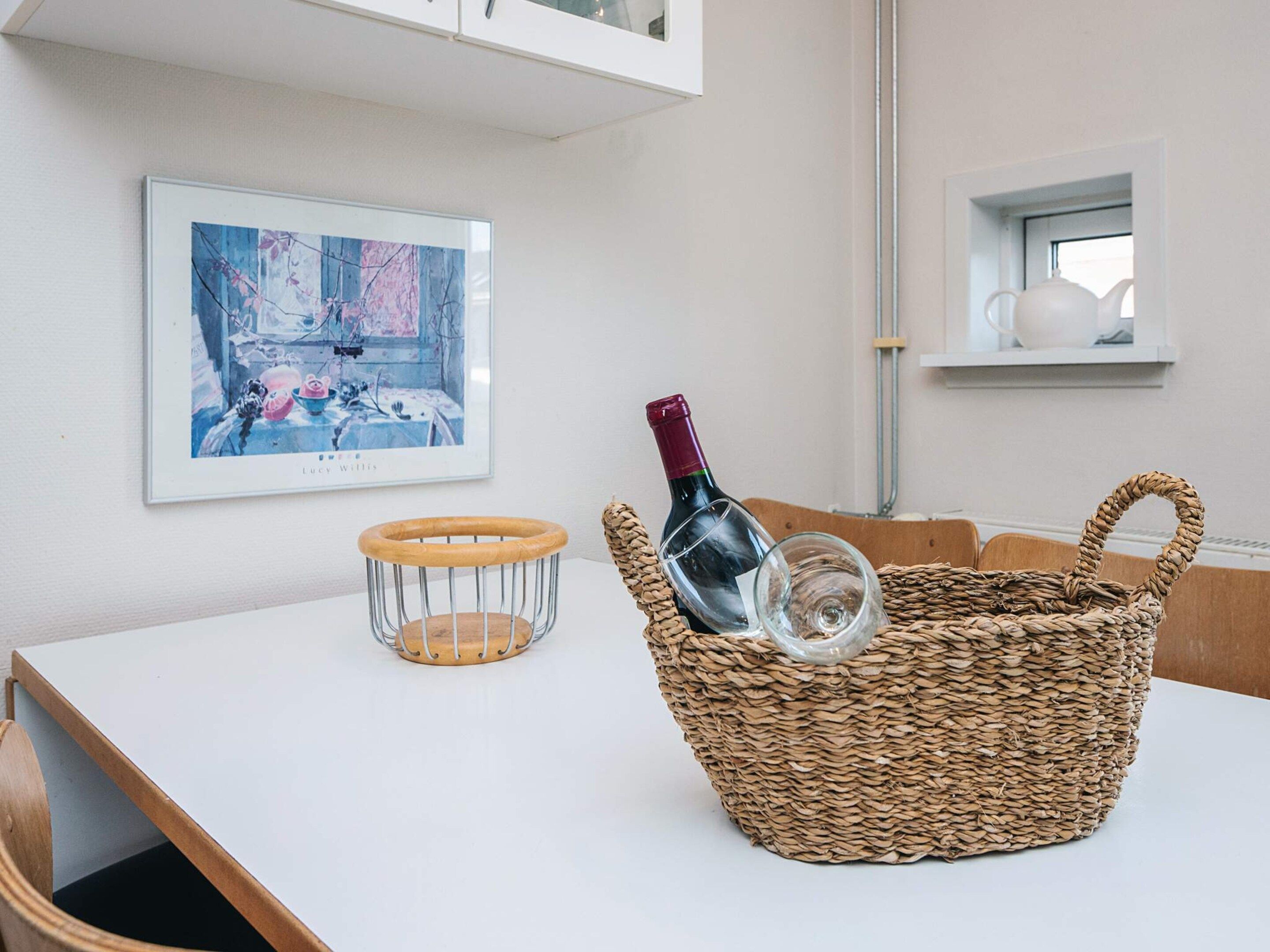 A dining area with a white table, a wicker basket with a wine bottle, a metal wine rack, and a framed picture on the wall.