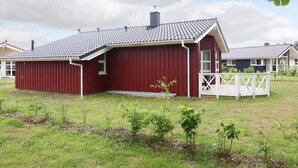 Un bâtiment rouge avec un toit en pente, une clôture blanche et une cour verdoyante où poussent de jeunes plantes.