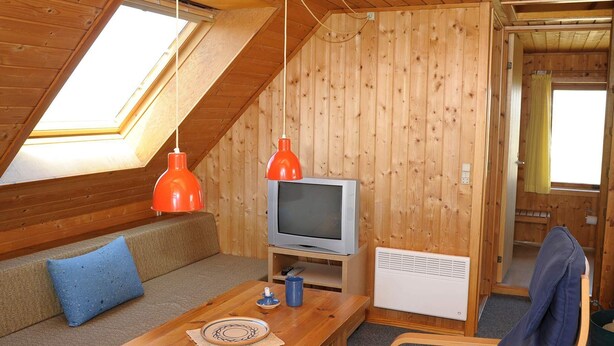 Ett mysigt rum på vinden med ett sluttande tak av trä, en tv, ett bord med en bordsduk, en soffa med kuddar och en stol.