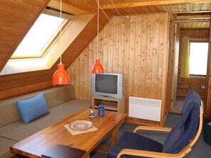 Ein gemütliches Dachzimmer mit Holzbalkendecke, einem Fernseher, einem Tisch mit Unterlage, einer Couch mit Kissen und einem Sessel.