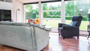 Un salon moderne avec un canapé gris, un fauteuil foncé et un téléviseur posé sur un meuble. De grandes fenêtres offrent une vue sur un jardin verdoyant.