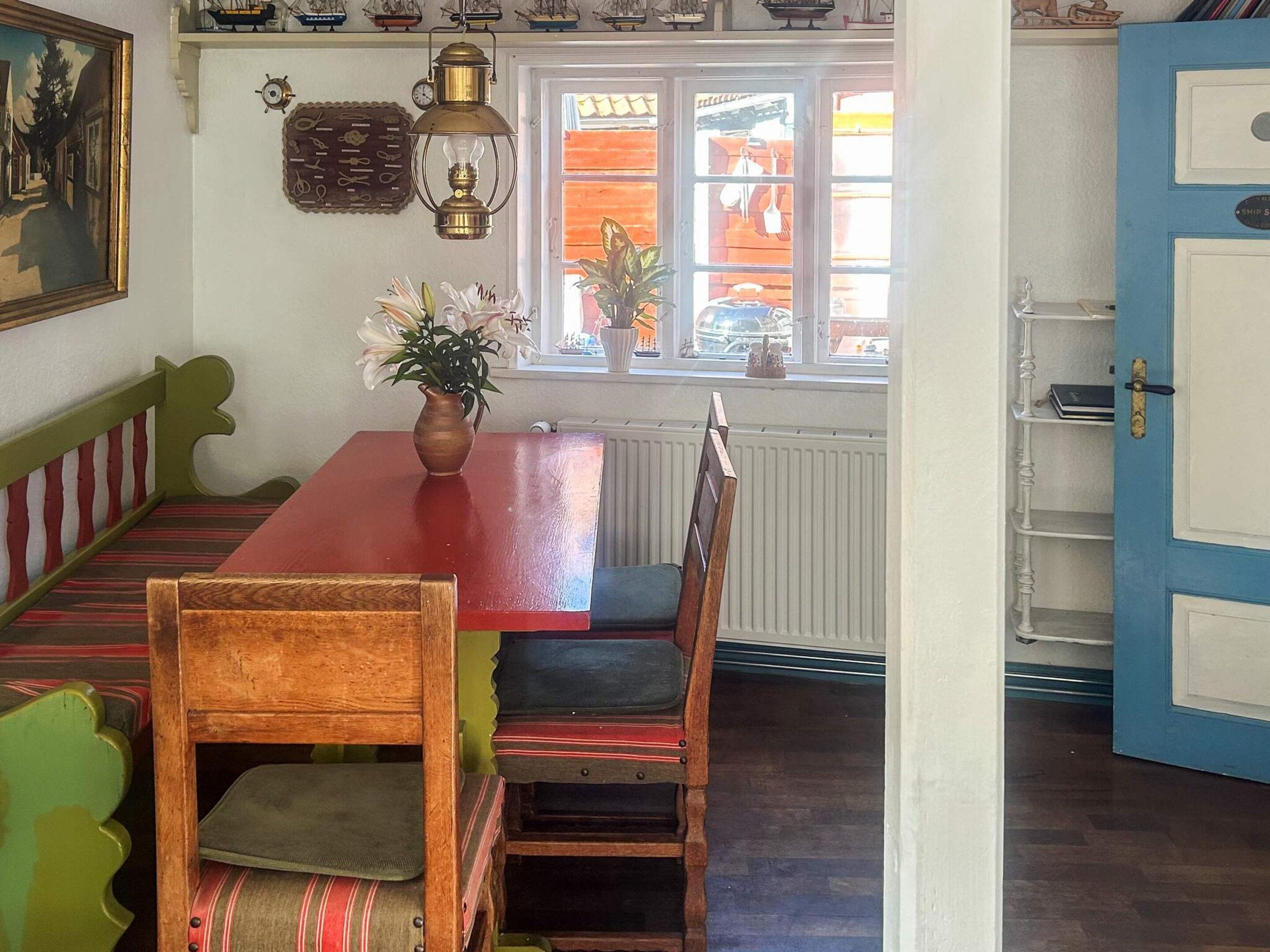 Un coin repas avec une table rouge, des chaises en bois et un banc. Il y a une fenêtre donnant sur la rue, et une porte sur la droite.