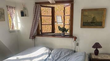 A bedroom with a bed, a window with curtains, a small table with a lamp, and a painting on the wall.