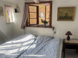 Un dormitorio con cama, ventana con cortinas, una mesita de noche con lámpara y un cuadro en la pared.