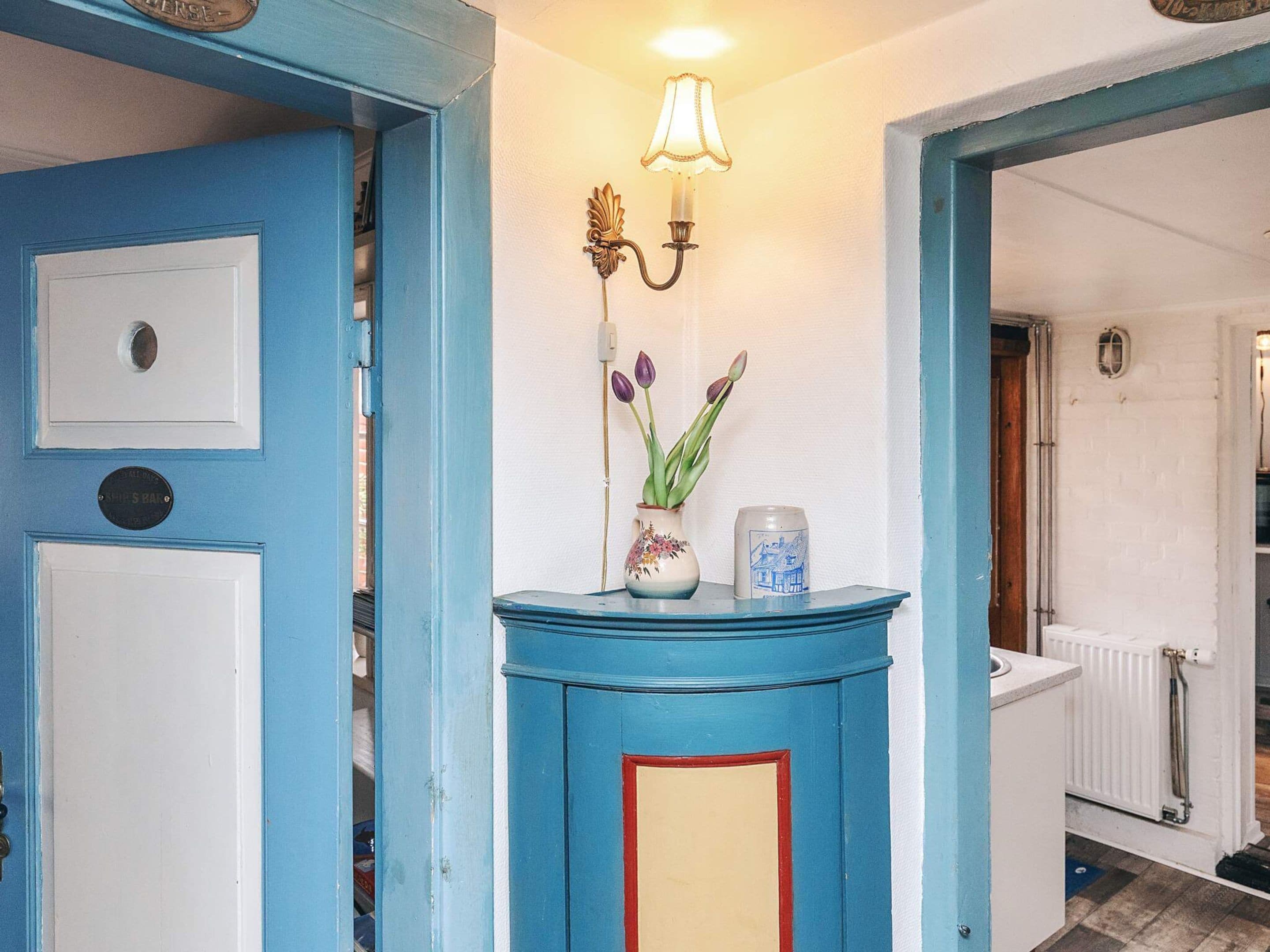 Una puerta azul con un picaporte blanco, una lámpara fijada a la pared y un jarrón con flores.