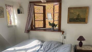 Un dormitorio con cama, una ventana con cortinas, una mesita de noche con lámpara y un cuadro en la pared.