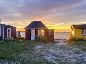 Escena costera con dos pequeñas casas de madera, una roja y otra amarilla, con vista despejada al mar y un atardecer de fondo.