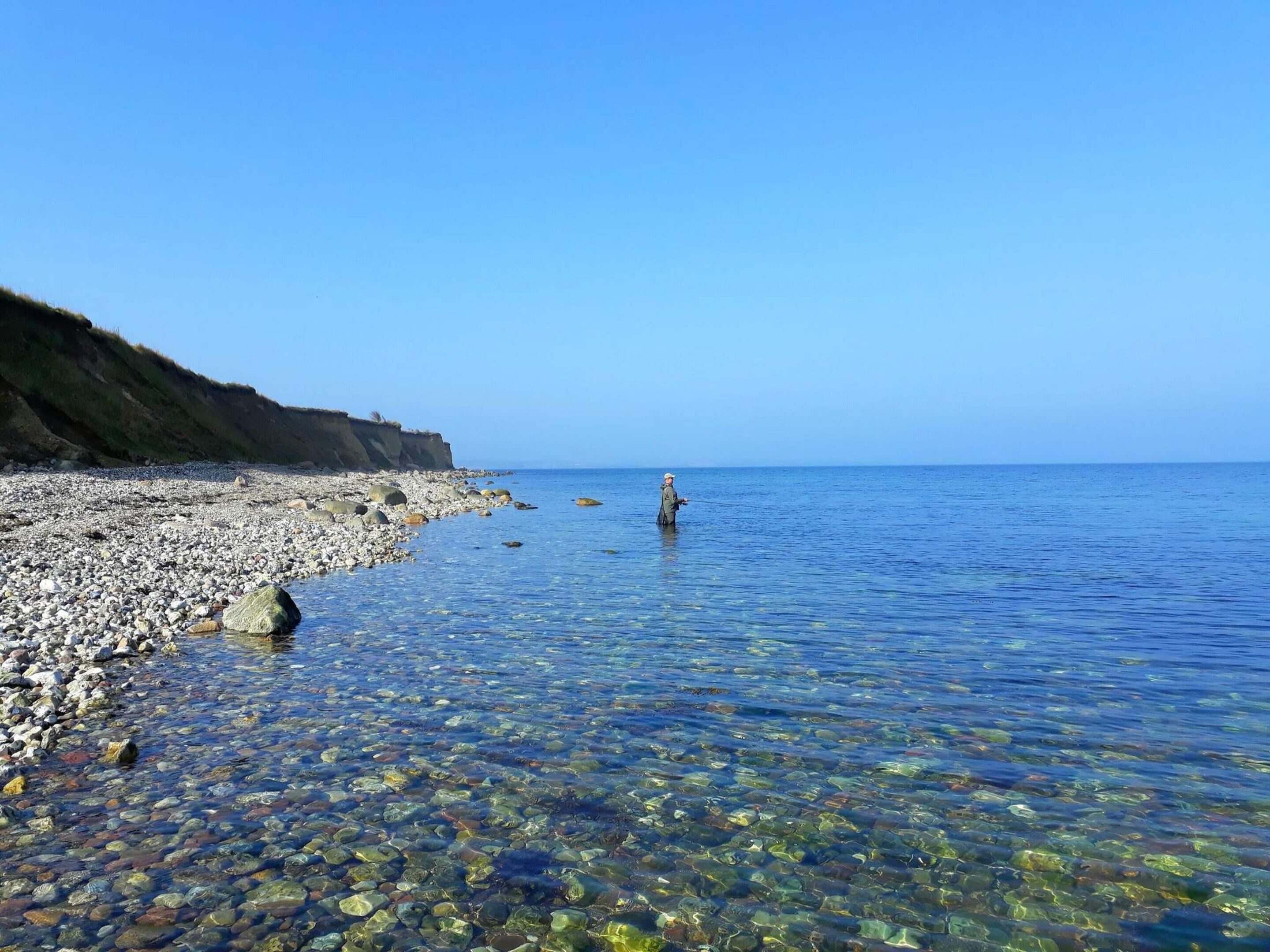 A clear beach with pebbles and a person standing in the water.
